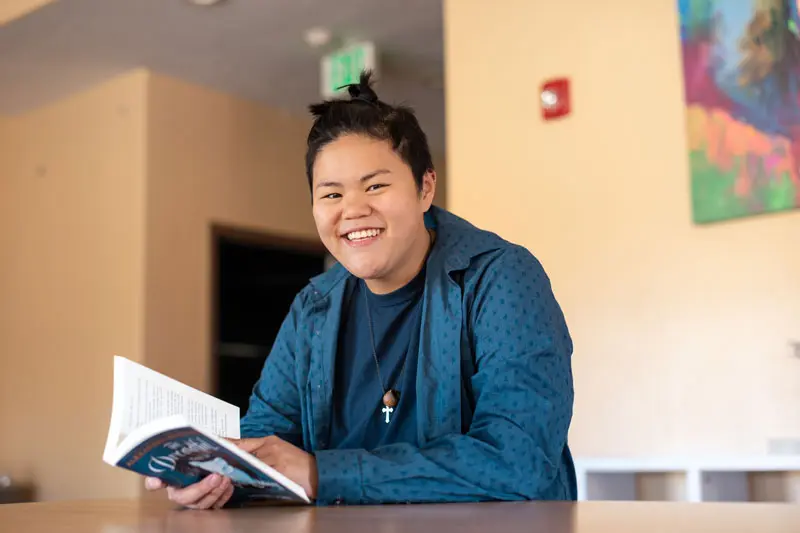 A student takes a break from reading a book to smile for the camera while attending a teen treatment center for girls and youth assigned female at birth | Discovery Ranch South